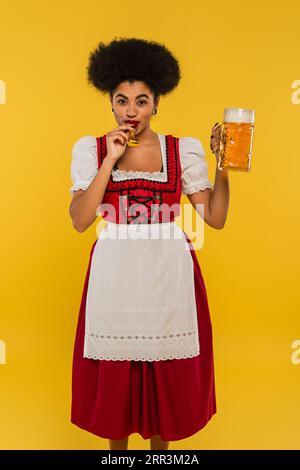 Fröhliche afroamerikanische oktoberfest-Kellnerin mit einem Becher Bier, der in Partyhorn auf Gelb bläst Stockfoto