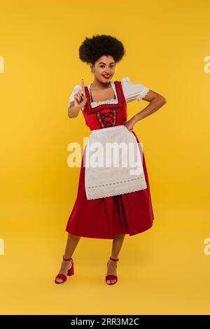 Fröhliche afroamerikanische oktoberfest-Kellnerin mit Hand auf Hüfte und gelbem Ideenschild Stockfoto