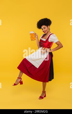 Fröhliche afroamerikanische Kellnerin im oktoberfest-Outfit mit Brezel und Bier auf gelbem Hintergrund Stockfoto