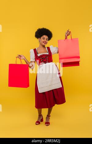 Fröhliche afroamerikanische bayerische Kellnerin in traditionellem Kleid, stehend mit Einkaufstaschen auf gelb Stockfoto