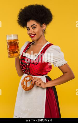 Charmante afroamerikanische oktoberfest-Kellnerin mit Bierbecher und Brezel lächelnd vor der Kamera auf Gelb Stockfoto