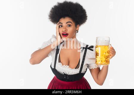 Beeindruckte afroamerikanische oktoberfest-Kellnerin mit Bierbecher und berührendem Gesicht auf weiß Stockfoto