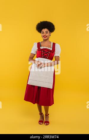 Fröhliche afroamerikanische bayerische Kellnerin im oktoberfestkostüm auf gelbem, durchgehendem Bein Stockfoto