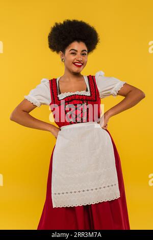 Fröhliche afroamerikanische bayerische Kellnerin im Dirndl, die Hände auf Taille auf gelbem oktoberfest hält Stockfoto