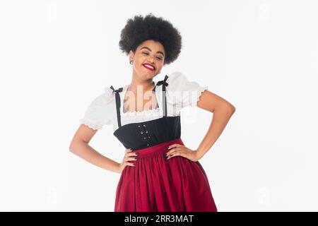 Fröhliche afroamerikanische Kellnerin in bayerischer Tracht mit Händen auf Hüften auf weiß, oktoberfest Stockfoto