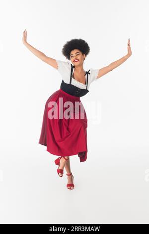 Fröhliche afroamerikanische bayerische Kellnerin in elegantem Dirndl mit oktoberfest-Tanz auf weiß Stockfoto