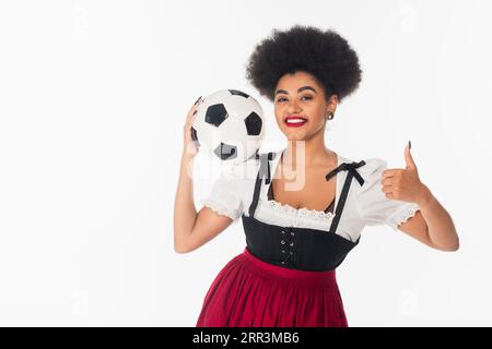 Fröhliche afroamerikanische oktoberfest-Kellnerin im Dirndl, die Fußball hält und wie auf weiß zeigt Stockfoto