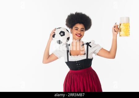 Fröhliche afroamerikanische Kellnerin im bayerischen Dirndl mit Fußballball und einem Becher Bier auf weiß Stockfoto