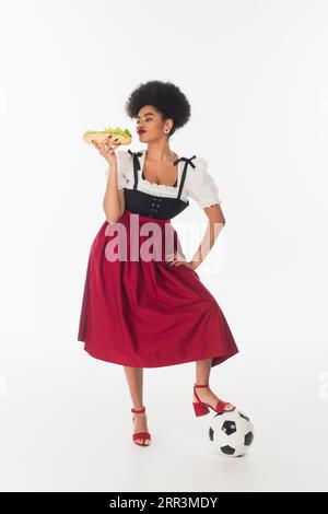 afroamerikanische bayerische Kellnerin posiert mit leckerem Hot Dog und Fußball auf weißem oktoberfest Stockfoto