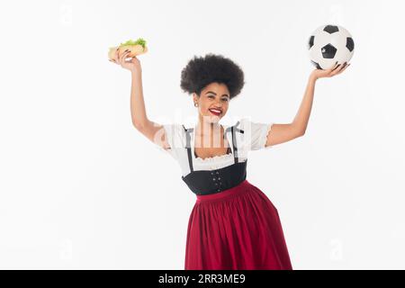 Begeisterte afroamerikanische bayerische Kellnerin mit Fußball und Hot Dog in erhobenen Händen auf weiß Stockfoto