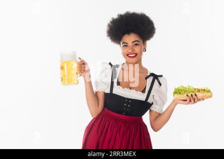 Fröhliche afroamerikanische bayerische Kellnerin mit Bierbecher und Hot Dog auf weißem oktoberfest-Konzept Stockfoto