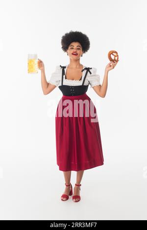 Fröhliche afroamerikanische bayerische Kellnerin im oktoberfest Kostüm mit Bierkrug und Brezel auf weiß Stockfoto