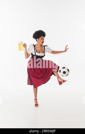 Fröhliche afroamerikanische bayerische Kellnerin mit einem Becher mit schaumigem Bären, der auf den Fußball auf weiß tritt Stockfoto