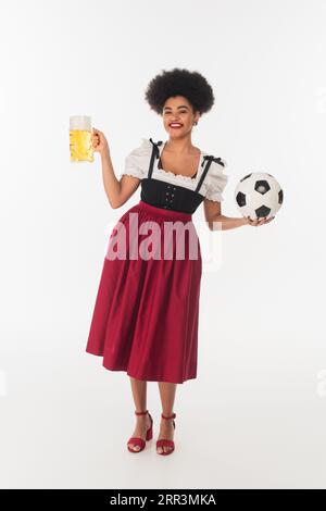 afroamerikanische bayerische Kellnerin posiert mit Fußball und Becher Craft-Bier mit Schaum auf weiß Stockfoto