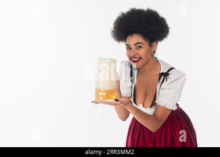 afroamerikanische oktoberfest-Kellnerin, die einen Becher Bier mit Schaum hält und die Kamera auf weiß schaut Stockfoto