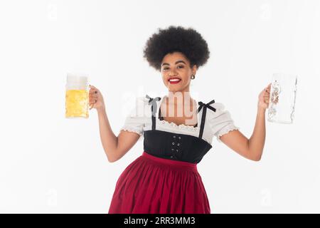 afroamerikanische bayerische Kellnerin im oktoberfest Dirndl mit leeren und vollen Bierbechern auf weiß Stockfoto