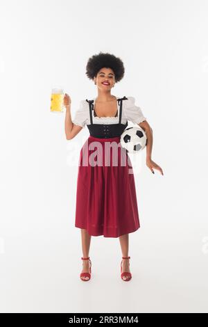 afroamerikanische bayerische Kellnerin mit Fußballball und einem Becher Bier, das auf weiß lächelt, oktoberfest Stockfoto