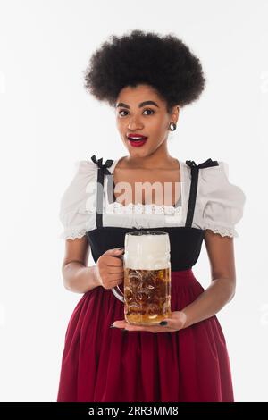 Staunende afroamerikanische bayerische Kellnerin im oktoberfest-Kostüm mit einem Becher schaumigem Bier auf weiß Stockfoto