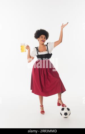 Fröhliche afroamerikanische oktoberfest-Kellnerin mit einem Becher Craft-Schaumbier in der Nähe des Fußballballs auf weiß Stockfoto