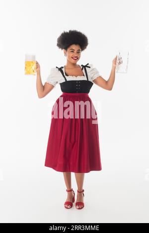 afroamerikanische oktoberfest-Kellnerin in traditioneller Tracht mit leeren und vollen Bierbechern auf weiß Stockfoto