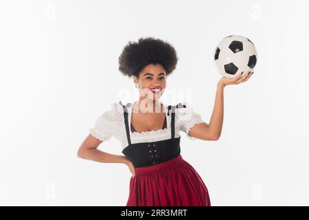 Lächelnde afroamerikanische bayerische Kellnerin im Dirndl mit Fußball auf weiß, oktoberfest-Konzept Stockfoto