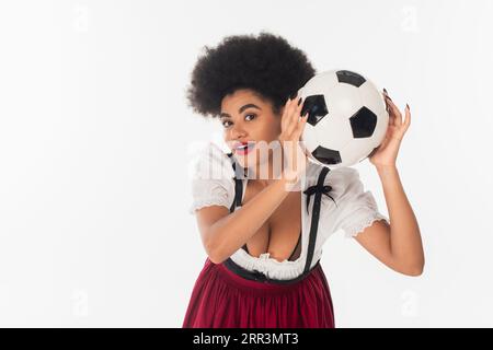 Aufgeregte afroamerikanische oktoberfest-Kellnerin mit Fußballball, der die Kamera auf weiß anschaut Stockfoto