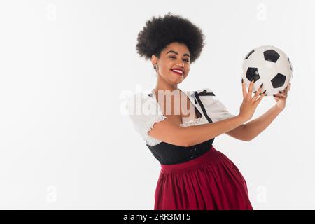 Fröhliche oktoberfest-Kellnerin in bayerischem Kostüm, die den Fußball afroamerikaner auf weiß hält Stockfoto