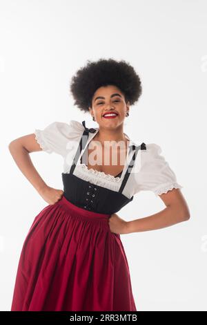 Fröhliche afroamerikanische oktoberfest-Kellnerin in traditionellem Dirndl-Kleid mit Händen auf Hüften auf weiß Stockfoto