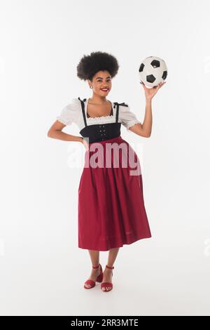 Verführerische afroamerikanische bayerische Kellnerin in authentischem Kleid, die mit Fußball auf weiß posiert Stockfoto