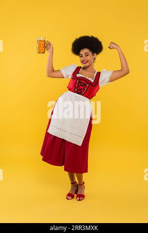 Aufgeregte afroamerikanische oktoberfest-Kellnerin mit Bierbecher mit Gewinngeste auf Gelb Stockfoto