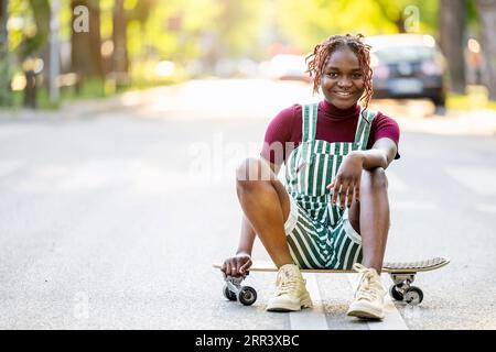 Porträt einer schwarzen, nicht binären Person mit einem Skateboard, das auf der Straße sitzt Stockfoto