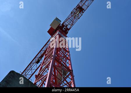 Ein Industrieturmkran von roter Farbe mit einem gestreckten Arm und einer weißen Fahrerkabine, die von unten beobachtet wird. Stockfoto