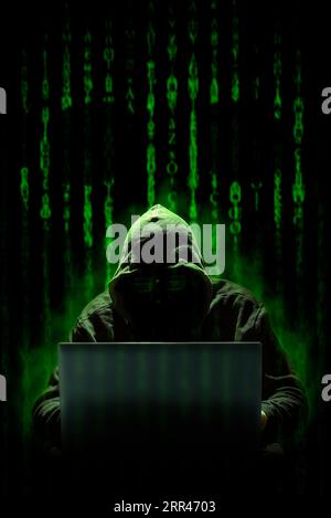 Ein Hacker mit Kapuze arbeitet mit einem Laptop an einem Cyberangriff, vor einem Hintergrund mit Computercode im Matrixstil, auf dem ein Schädel zu sehen ist. Cyberkriminalität. Stockfoto