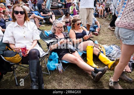 Zwei Festivalbesucher mittleren Alters, die in Gummistiefeln im Schlamm sitzen und auf ihren Mobiltelefonen scrollen. Green man Festival, Brecon, Wales, Großbritannien, 2023. Foto: Rob Stockfoto