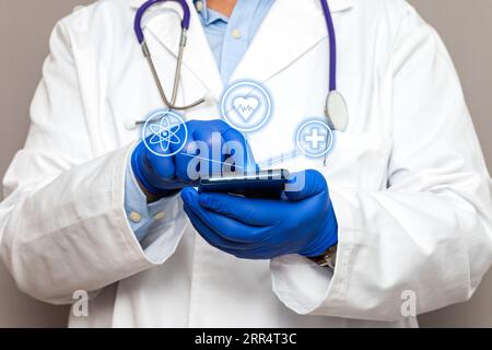 Nahaufnahme der Hände eines nicht erkennbaren Arztes, der blaue Handschuhe trägt und ein Smartphone hält, während er den Geröll berührt und drei Symbole schweben. Stockfoto