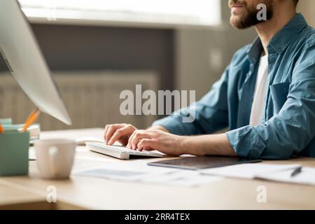 Bärtiger junger Mann, der am Schreibtisch sitzt und auf der Computertastatur schreibt Stockfoto