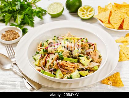 Thunfischsalat mit Avocado, roter Zwiebel, Gurke und Kapern mit Olivenöl, Limetten-Petersilie-Dressing in weißer Schüssel auf weißem Holztisch mit Taco-Chips und f Stockfoto