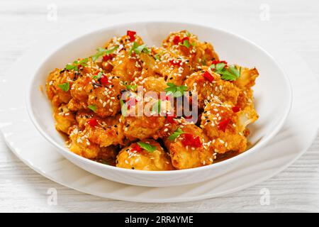 Knusprige orangefarbene Blumenkohlröschen, bestreut mit Sesamsamen, Chiliflocken und Petersilie in weißer Schüssel auf weißem Holztisch Stockfoto