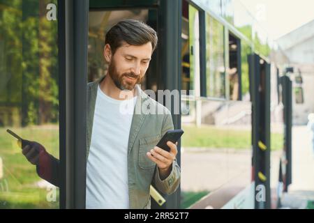 Eleganter junger Mann mit Smartphone und Ausstieg aus dem Bus in der Stadt. Vorderansicht des Bärtigen, der die Zeit auf dem Handy überprüft, während er den Transport an der Bushaltestelle verlässt. Transport, Planungskonzept. Stockfoto