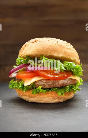 Köstlicher Burger mit saftigen Rindfleischschnitzeln, Käse, Tomaten, Salat, Zwiebeln auf einer schwarzen Steinplatte auf dunklem Holzhintergrund. Fast Food, hausgemachter Schinken Stockfoto