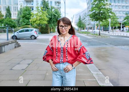 Reife lächelnde Frau in gesticktem Hemd mit Blick auf die Kamera, auf der Straße der modernen Stadt Stockfoto