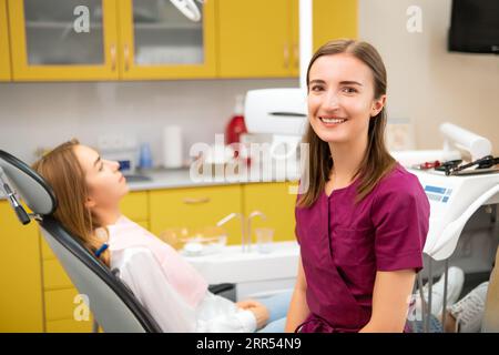 Zahnärztin schaut auf die Kamera, während Patient auf die Prozedur wartet und auf dem medizinischen Stuhl sitzt Stockfoto