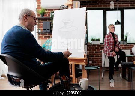 Seniormann, der an der Staffelei sitzt, Zeichnung übt, Gruppenkunstkurse besucht, im Ruhestand Neues lernt. Reife männliche Studenten skizzieren mit Bleistift auf Leinwand und besuchen kreative Kunstaktivitäten Stockfoto