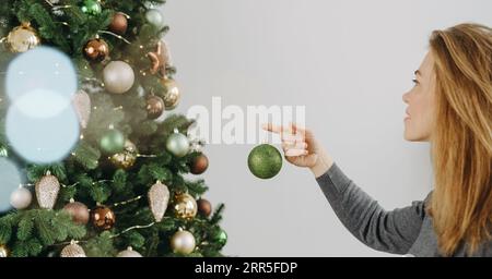 Frau, die den großen Weihnachtsbaum dekoriert. Weibchen mittleren Alters, das eine grüne Kugel in der Hand hält und am Baumzweig hängt. Leerzeichen kopieren. Grußkarte. Banner Stockfoto