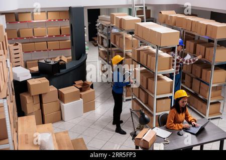 Das Team der Lagermitarbeiter arbeitet im Auftragsabwicklungszentrum. Asiatische Lagerarbeiter scannen Pakete, durchsuchen Pakete und führen Inventare auf der Draufsicht des Laptops durch Stockfoto