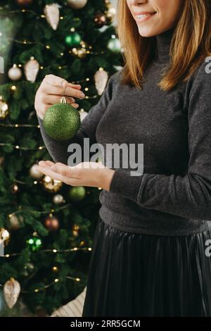 Frau, die den großen Weihnachtsbaum dekoriert. Weibchen mittleren Alters, das eine grüne Kugel in der Hand hält und am Baumzweig hängt. Leerzeichen kopieren. Grußkarte. Nahaufnahme Stockfoto