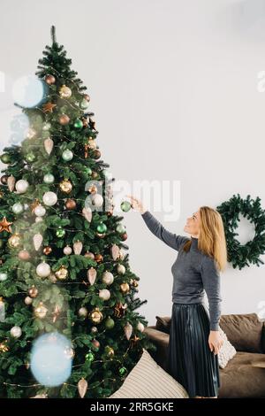Frau, die den großen Weihnachtsbaum dekoriert. Weibchen mittleren Alters, das eine grüne Kugel in der Hand hält und am Baumzweig hängt. Leerzeichen kopieren. Grußkarte. Stockfoto