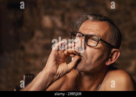 Ein älterer Mann, etwa 60 Jahre alt, serbischer Nationalität mit langen Haaren und Brille, raucht nachdenklich eine Zigarette auf dunklem Hintergrund. Künstler, cre Stockfoto