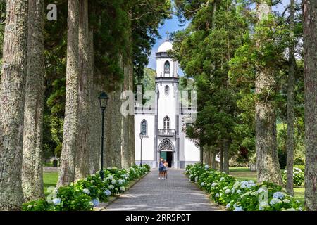 Junge Frauen nehmen ein Selfie vor der Igreja de São Nicolau oder der Kirche St. Nikolaus, die von Platanen und blühenden Hortensiensträuchern eingerahmt ist, im historischen Dorf Sete Cidades, Sao Miguel, Azoren, Portugal. Die 1857 erbaute Kirche befindet sich in der Mitte eines gewaltigen Vulkankraters, der drei Meilen (4,8 km) entfernt liegt. Stockfoto