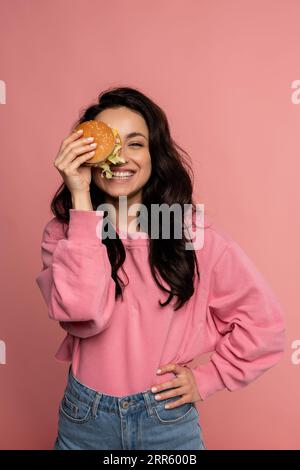 Lächelnde glückliche dunkelhaarige junge Frau, die mit ihrem köstlichen Sandwich vor der Kamera spielt. Appetit und beliebtes Fast-Food-Konzept Stockfoto